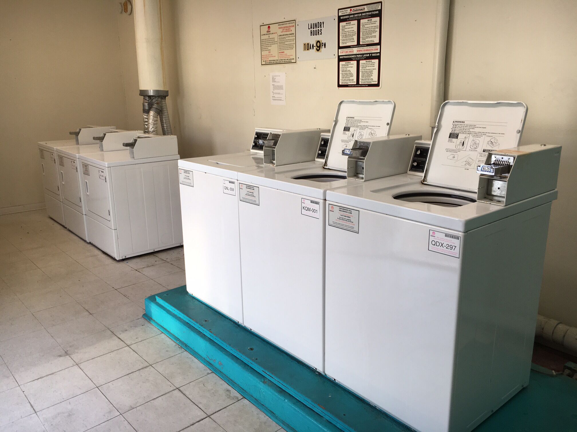A laundry facility with two washing machines and a dryer, a blue safety mat, and informational posters on the wall.