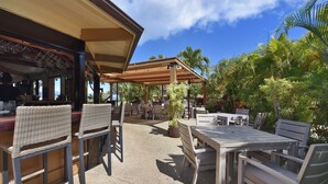 Un espace extérieur aménagé en terrasse, avec des chaises et des tables en osier, entouré de palmiers et offrant une vue sur l’océan.