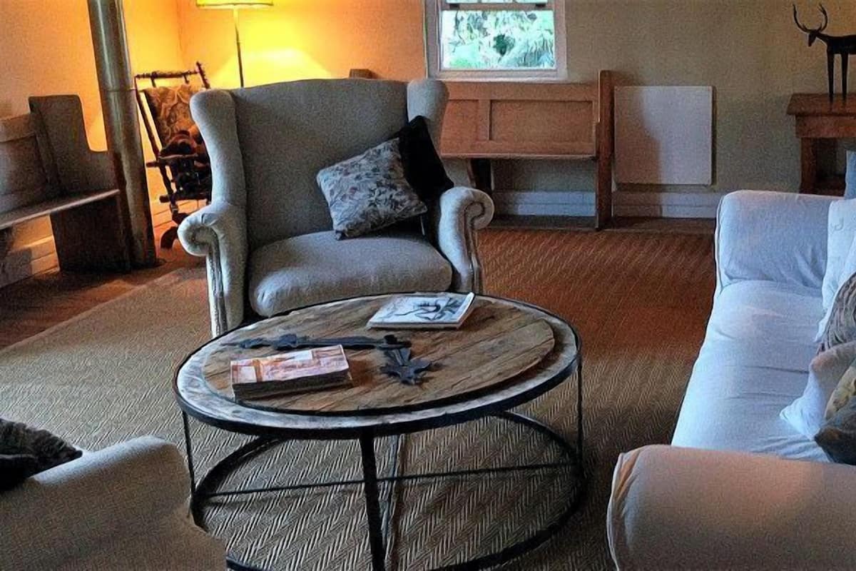 Una sala de estar acogedora con un sillón gris, un sofá blanco y una mesa redonda de centro con libros y una lámpara.