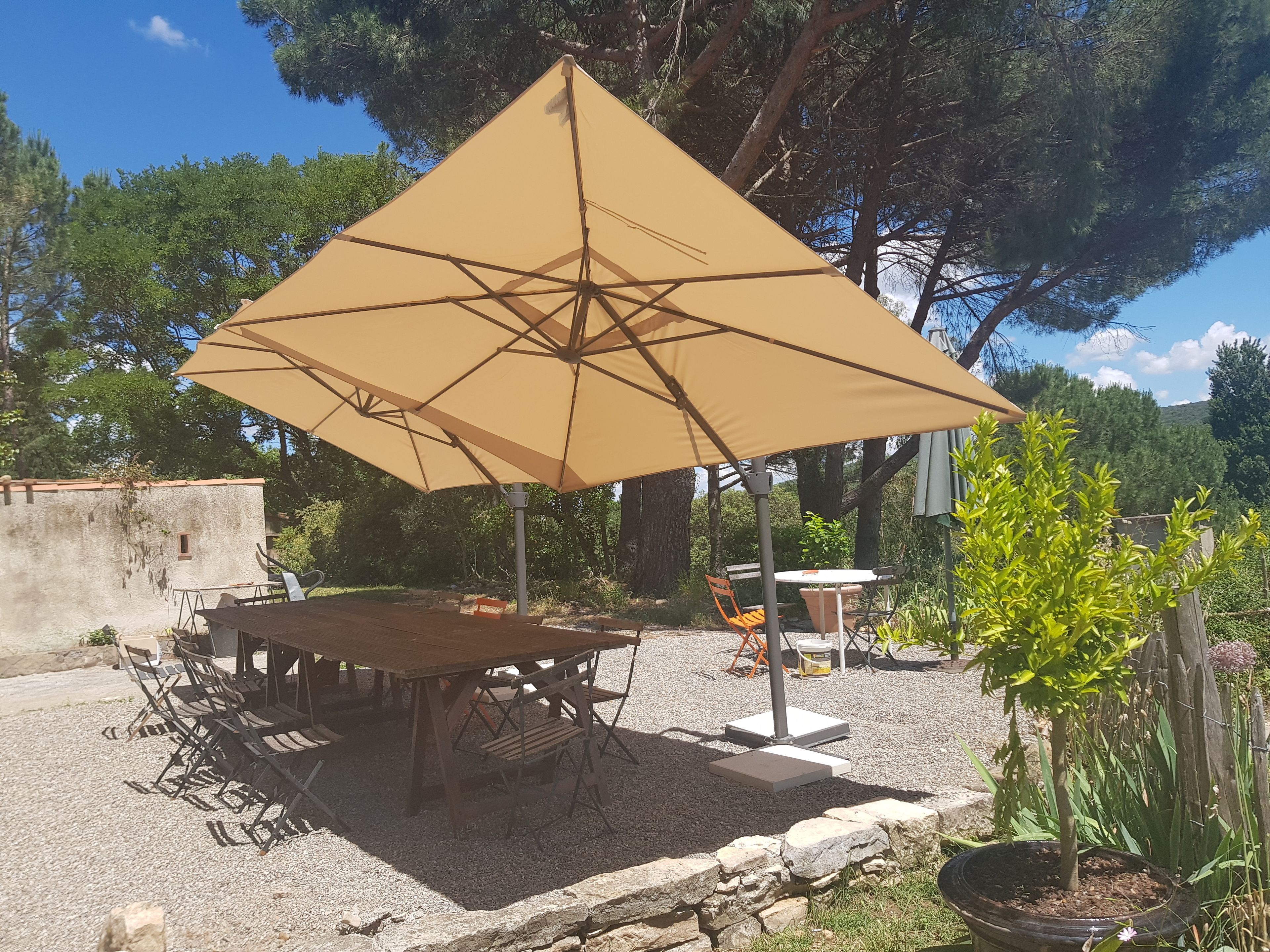 Een terras met een houten tafel en stoelen onder een grote parasol, omringd door grind en beplanting.