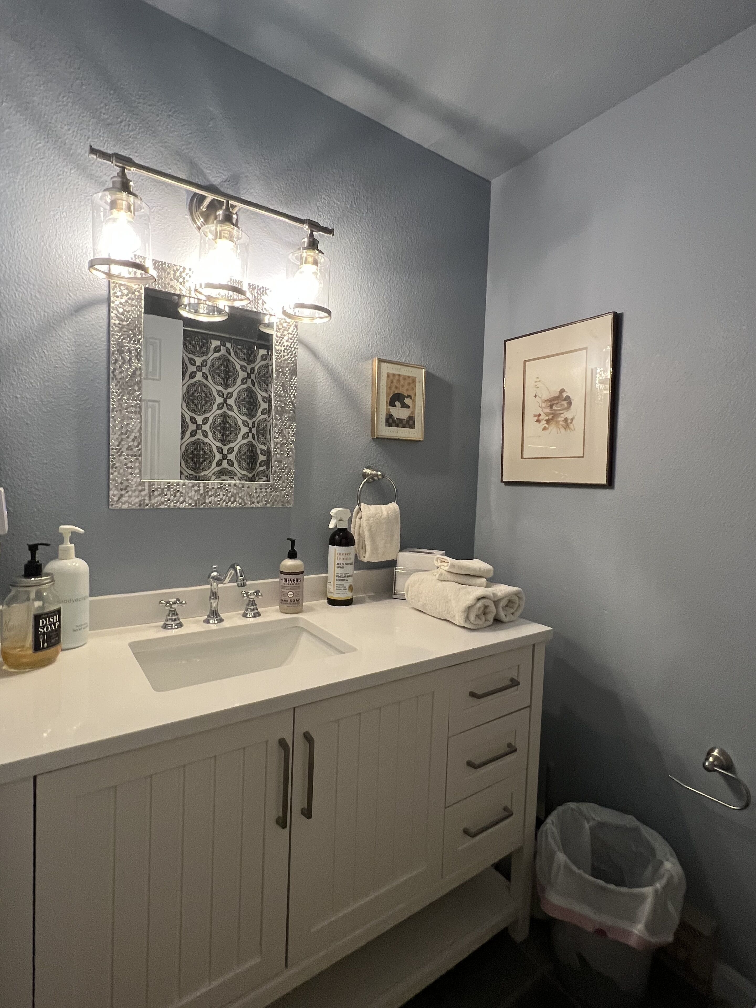A bathroom with a white vanity, a large mirror, and a sink. There are toiletries on the counter, a towel rack, and a trash can.