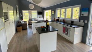 A modern kitchen with a central island, white cabinetry, and a large window providing natural light. -  Comfort and relaxation at Kawau Bay Beach House.  (Snells Beach)