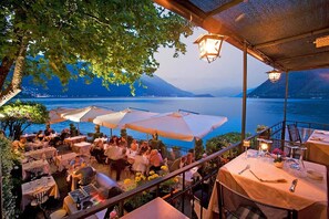 Un área de comedor al aire libre con mesas y sombrillas con vista a un cuerpo de agua y montañas.