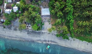 An aerial view of a coastal area with a beach, a small boat, and several buildings surrounded by palm trees.