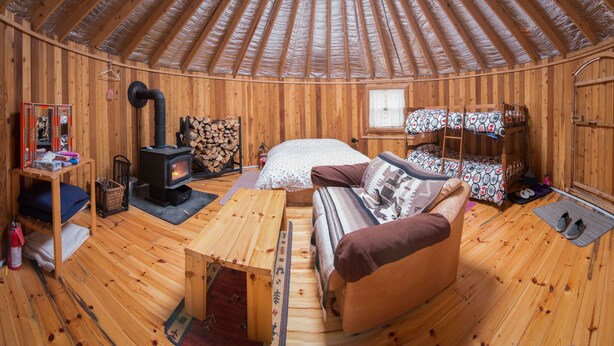 A cozy wooden interior with a sofa, a small table, a bed with patterned bedding, a wooden wardrobe, and a log storage area.