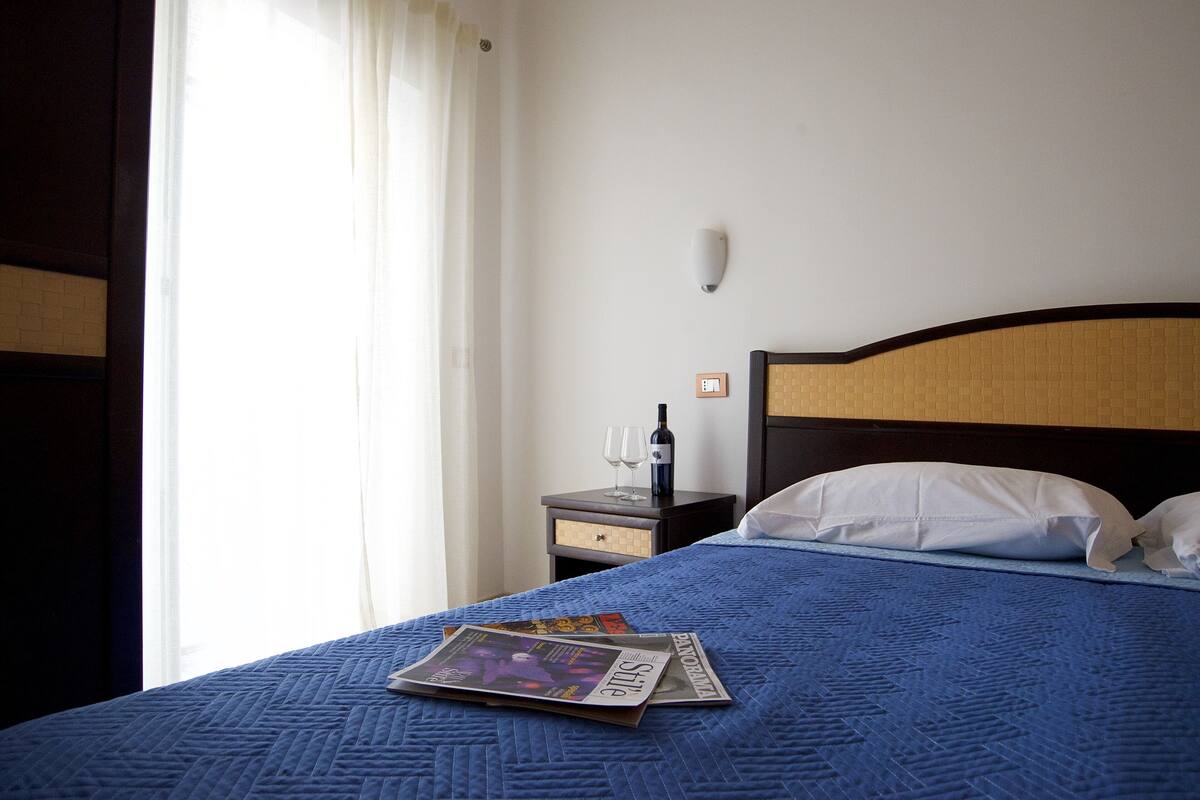 Een hotelkamer met een bed, nachtkastje, fles wijn en een tijdschrift.