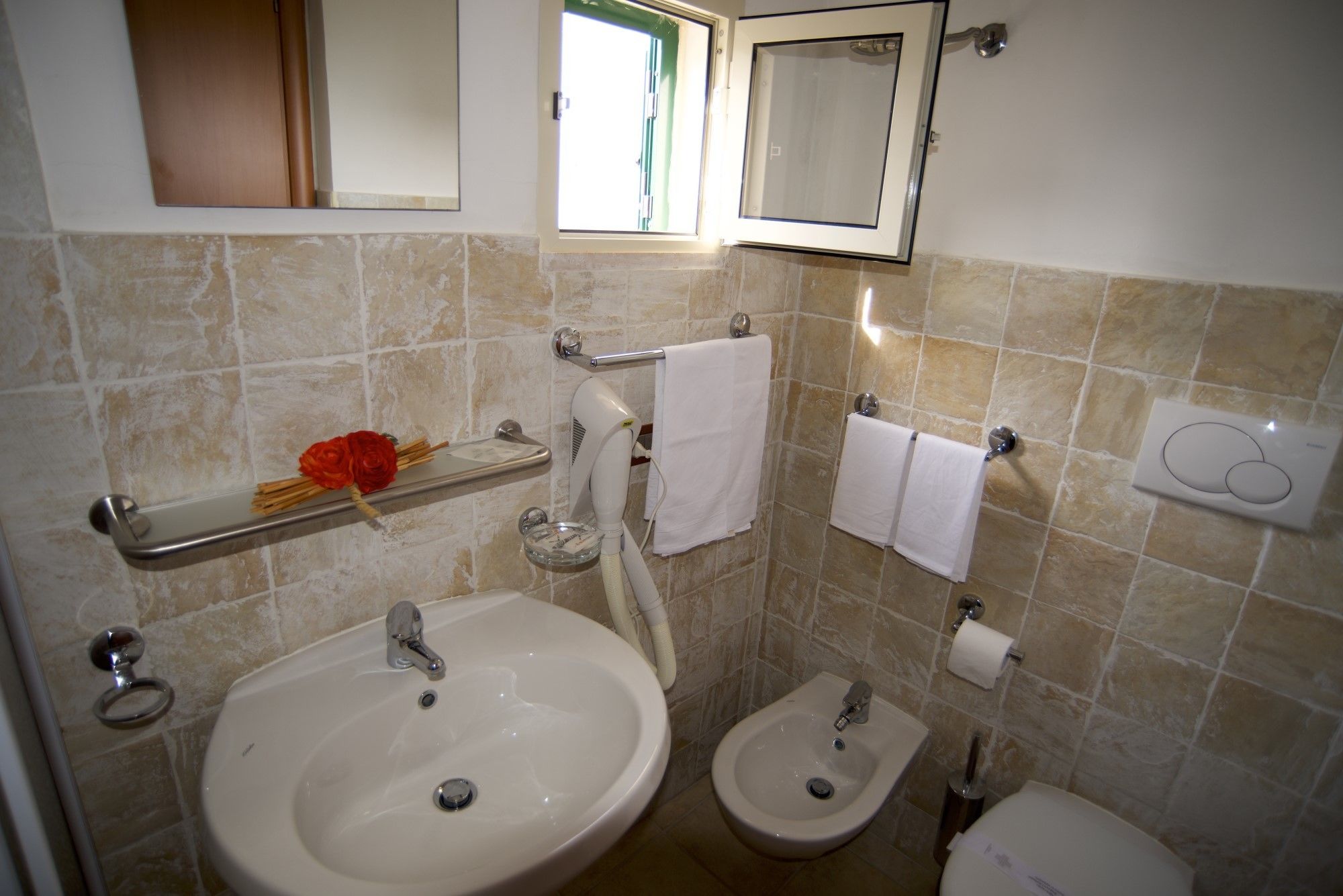A bathroom with a sink, bidet, and toilet, featuring beige tiles and a mirror above the sink.