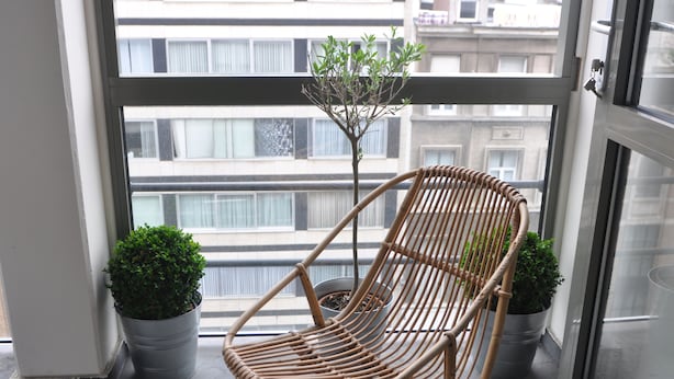 Una sedia in vimini con una pianta in vaso su un balcone che si affaccia su edifici.