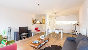 Un soggiorno moderno con un divano, un tavolino e una TV. È presente un'area pranzo con tavolo e sedie. In secondo piano si intravede la cucina con mobili bianchi.