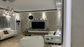 Un salon moderne avec un téléviseur à écran plat, des canapés blancs, une table basse et une horloge décorative au mur.