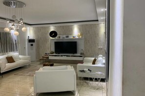 A modern living room with a flat-screen TV, white sofas, a coffee table, and a decorative clock on the wall. - Résidence 8 Tranche  T3 (Abidjan)