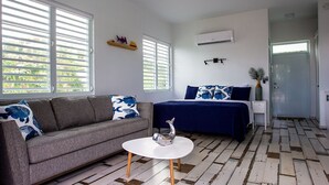 Um quarto com um sofá cinzento, uma cama com roupa de cama azul, uma mesa branca e uma almofada estampada.