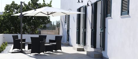 An outdoor patio with a table and chairs, a white umbrella, and a building with shutters.