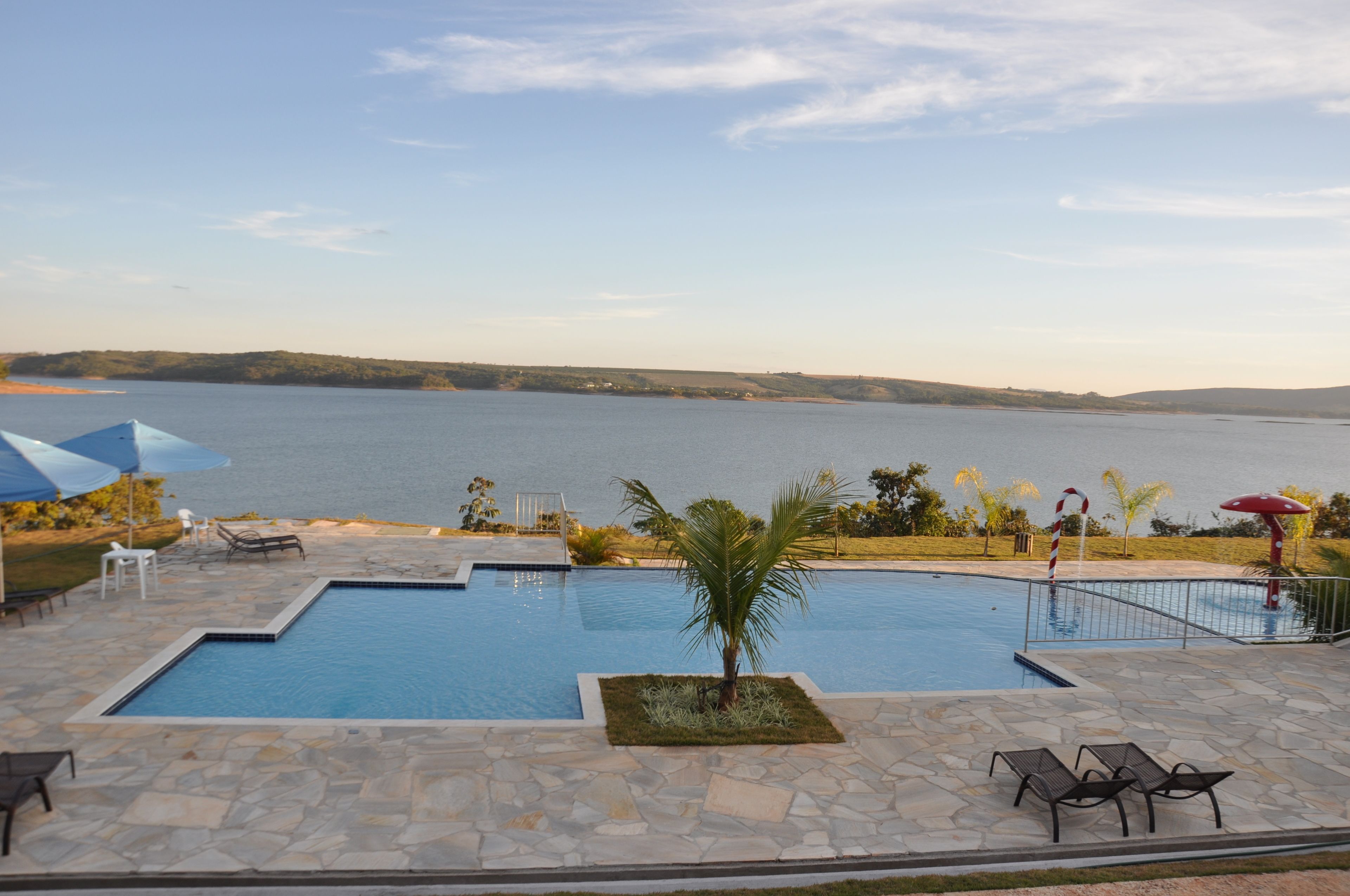 A swimming pool with a view of a lake, surrounded by stone tiles and equipped with lounge chairs and umbrellas.