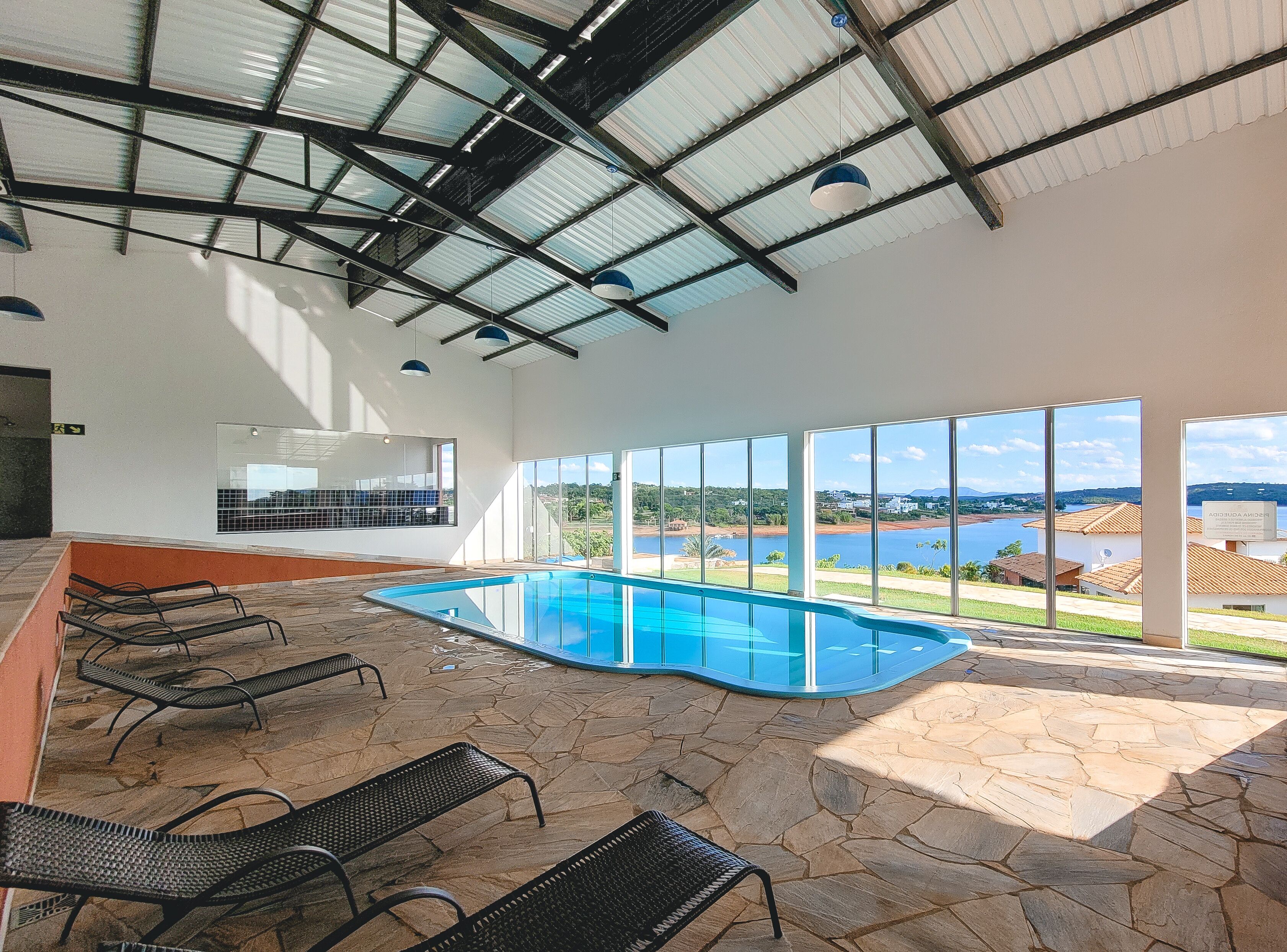 Una piscina con suelo de piedra y tumbonas de metal, ventanas amplias con vistas a un lago y edificios, y un techo alto con vigas de metal.