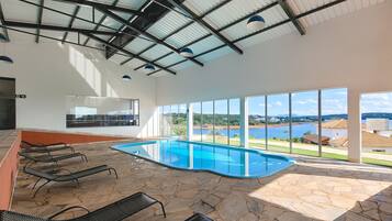 Una piscina con suelo de piedra y tumbonas de metal, ventanas amplias con vistas a un lago y edificios, y un techo alto con vigas de metal.