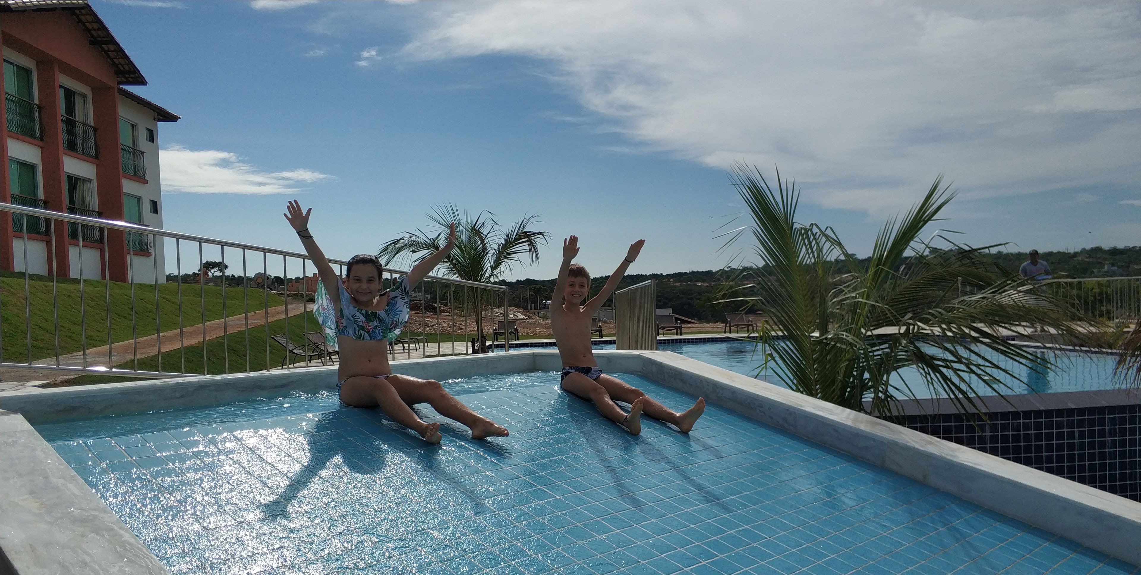 Uma piscina com duas pessoas sentadas na borda, um edifício com varandas e palmeiras ao fundo.