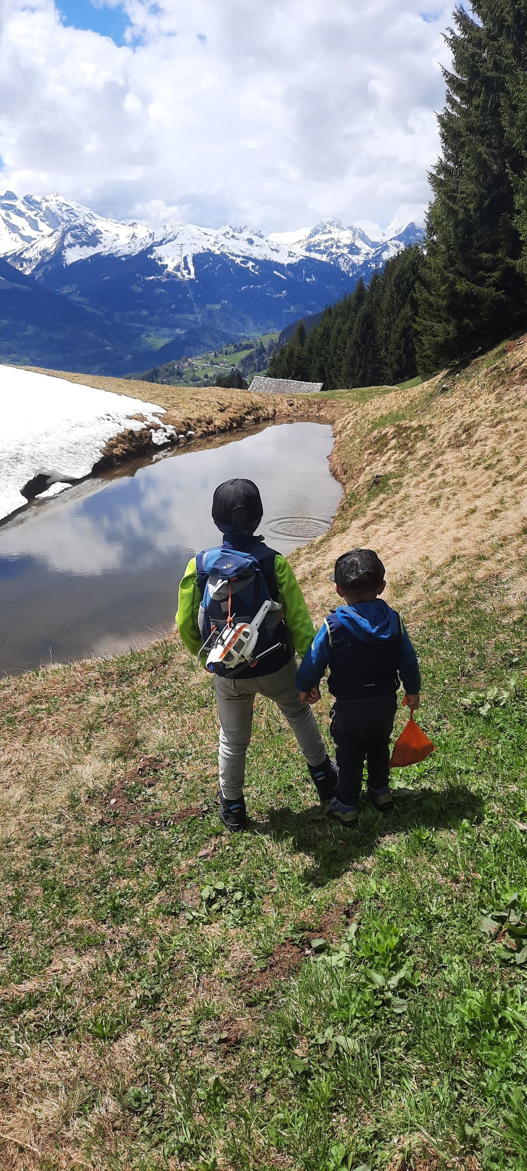Two children walking along a grassy path beside a calm lake with snow-capped mountains in the background.