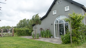 A grey building with a large arched window and a door, surrounded by greenery and a wooden gate.