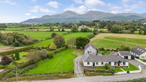 A rural landscape with a white house featuring a red door, surrounded by green fields and a mountain range in the background. - Ballyginny Cottage (Castlewellan)