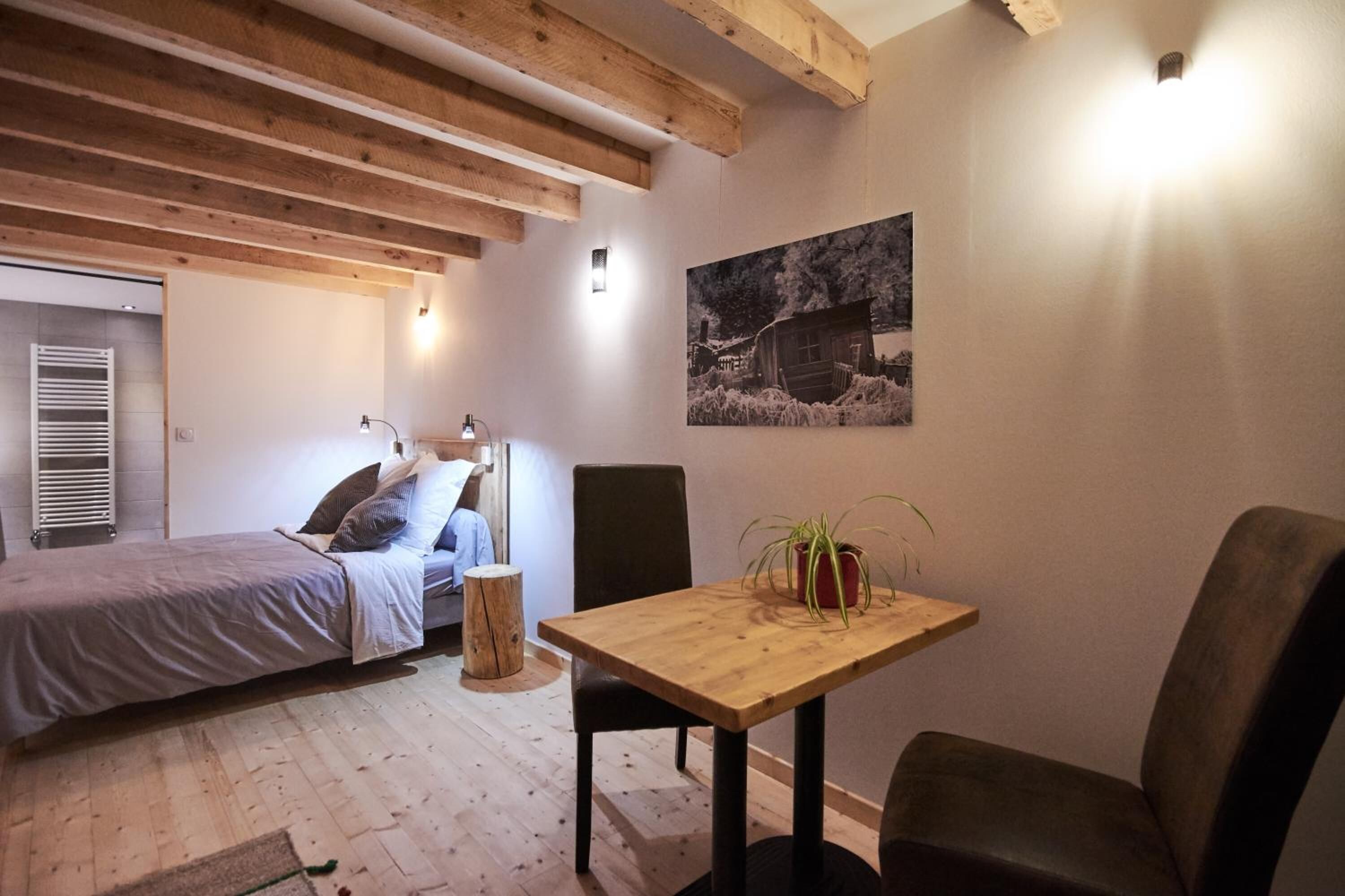 A cozy room with a bed, a wooden table with chairs, a small stool, and a wall-mounted light.