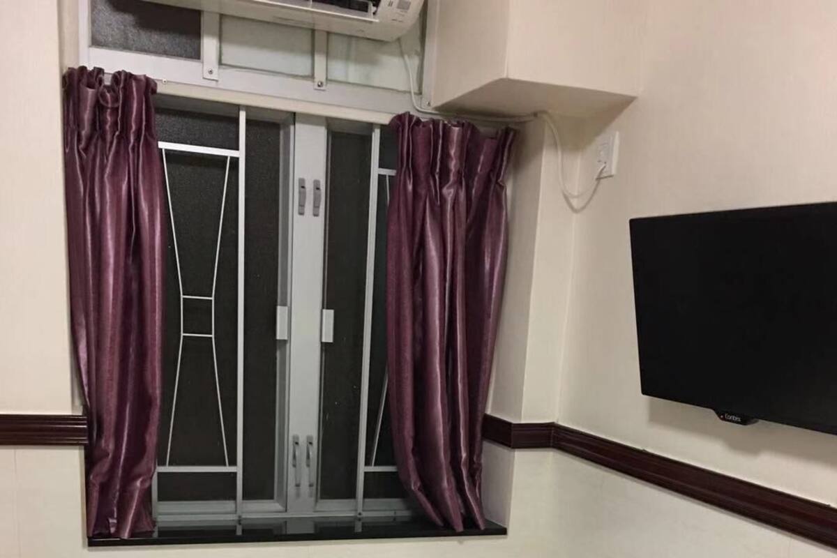 A hotel room with a bed, a window with purple curtains, a television mounted on the wall, and a small air conditioning unit above the window.