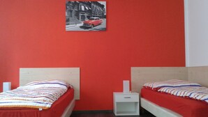 Een kamer met twee bedden, elk voorzien van een rode matras en gestreepte beddengoed. Daartussen staat een klein wit nachtkastje. De muren zijn rood geschilderd, en er hangt een ingelijst kunstwerk boven de bedden.