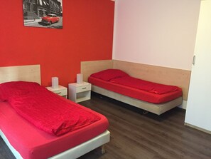 Een kamer met twee enkele bedden, elk met een rode matras en houten hoofdborden. Er zijn witte nachtkastjes met lampen en een klein ladeblok. De muren zijn rood en er hangt een ingelijst kunstwerk aan de muur.