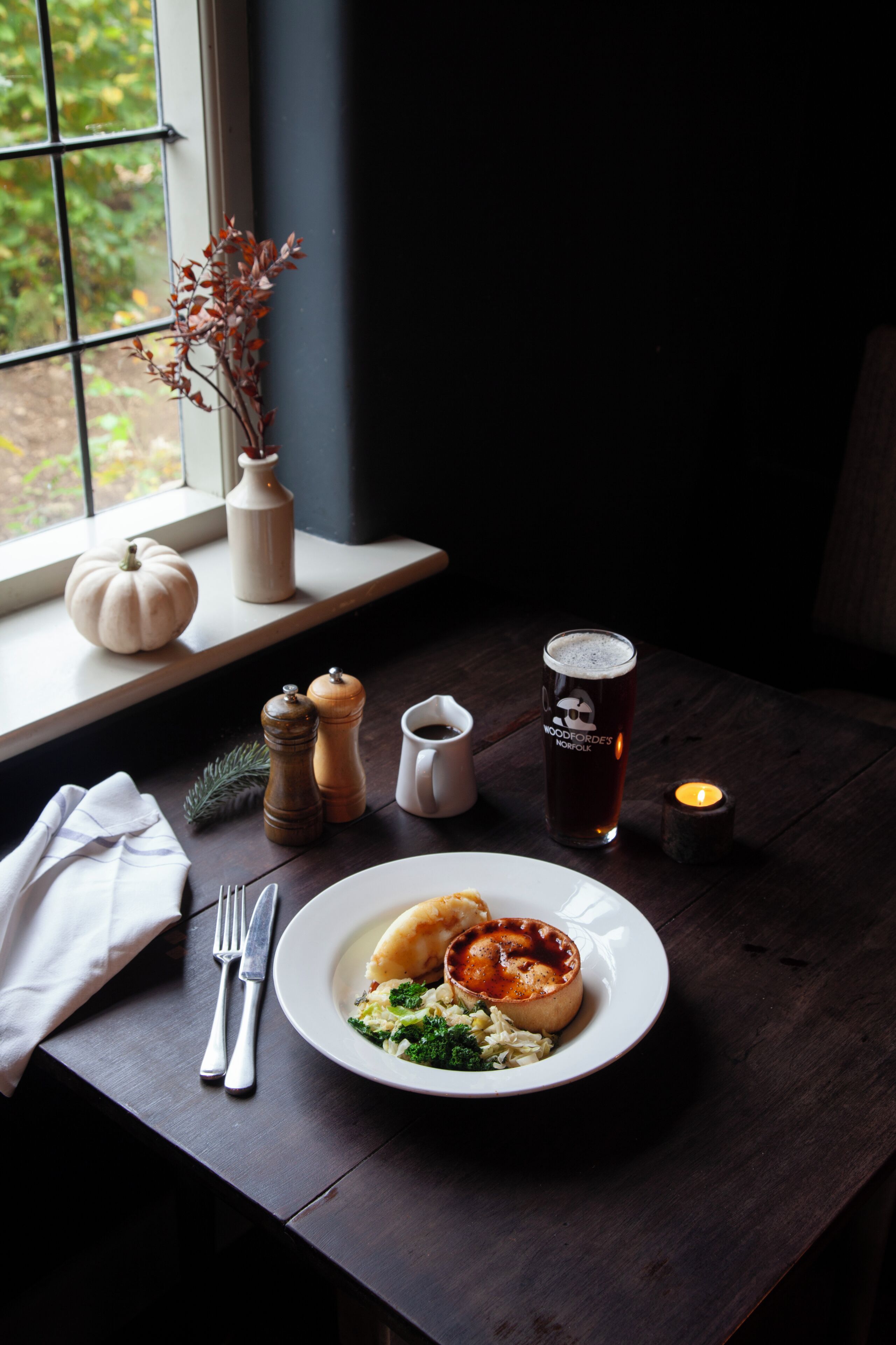 A dining table with a plate of food, a glass of beer, salt and pepper shakers, a candle, and a vase with dried branches.