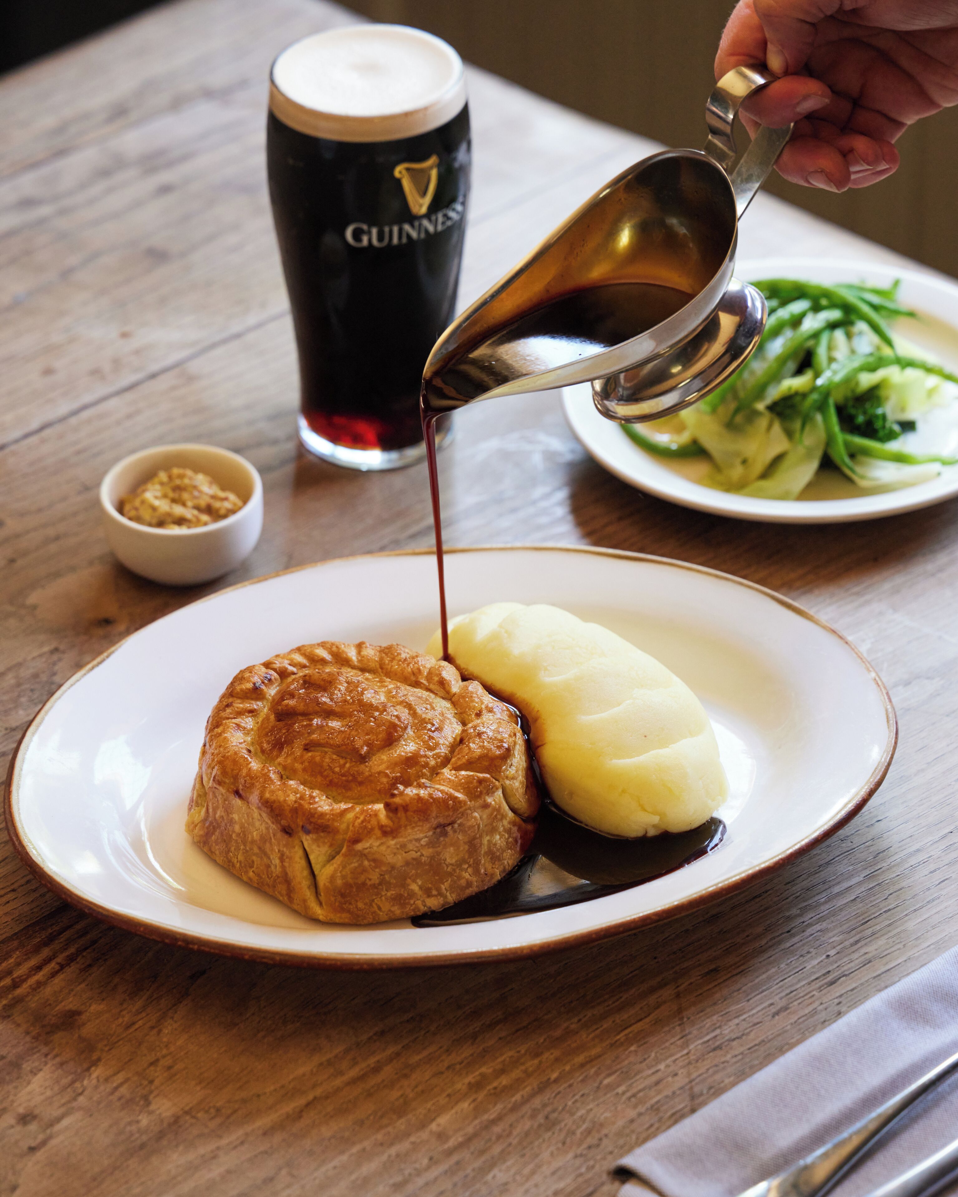 Une assiette avec une pâtisserie et de la purée de pommes de terre, un verre de bière Guinness et un petit bol de condiments sur une table en bois.