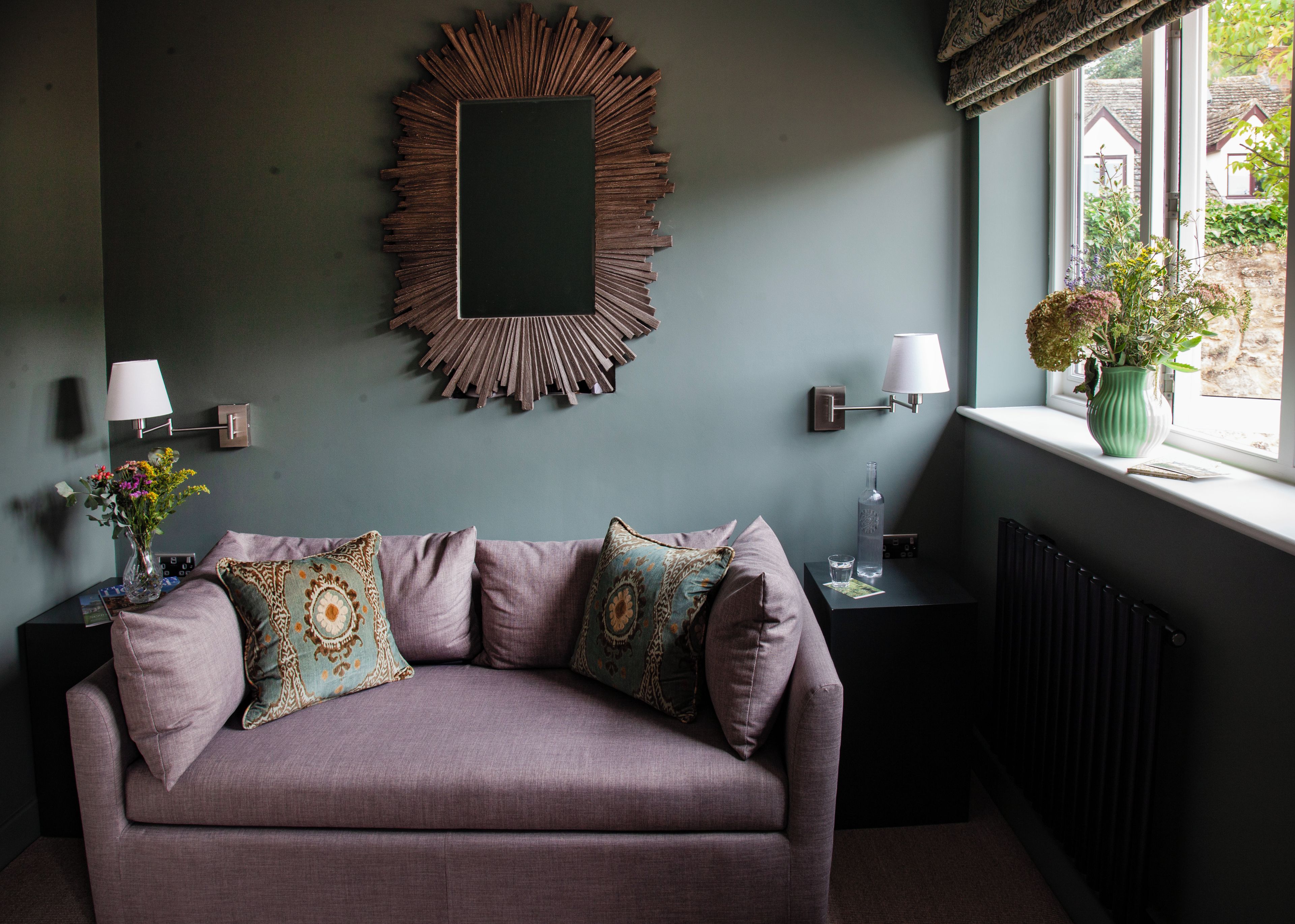 A room with a sofa, decorative pillows, wall-mounted lamps, a mirror, and a window with a view.