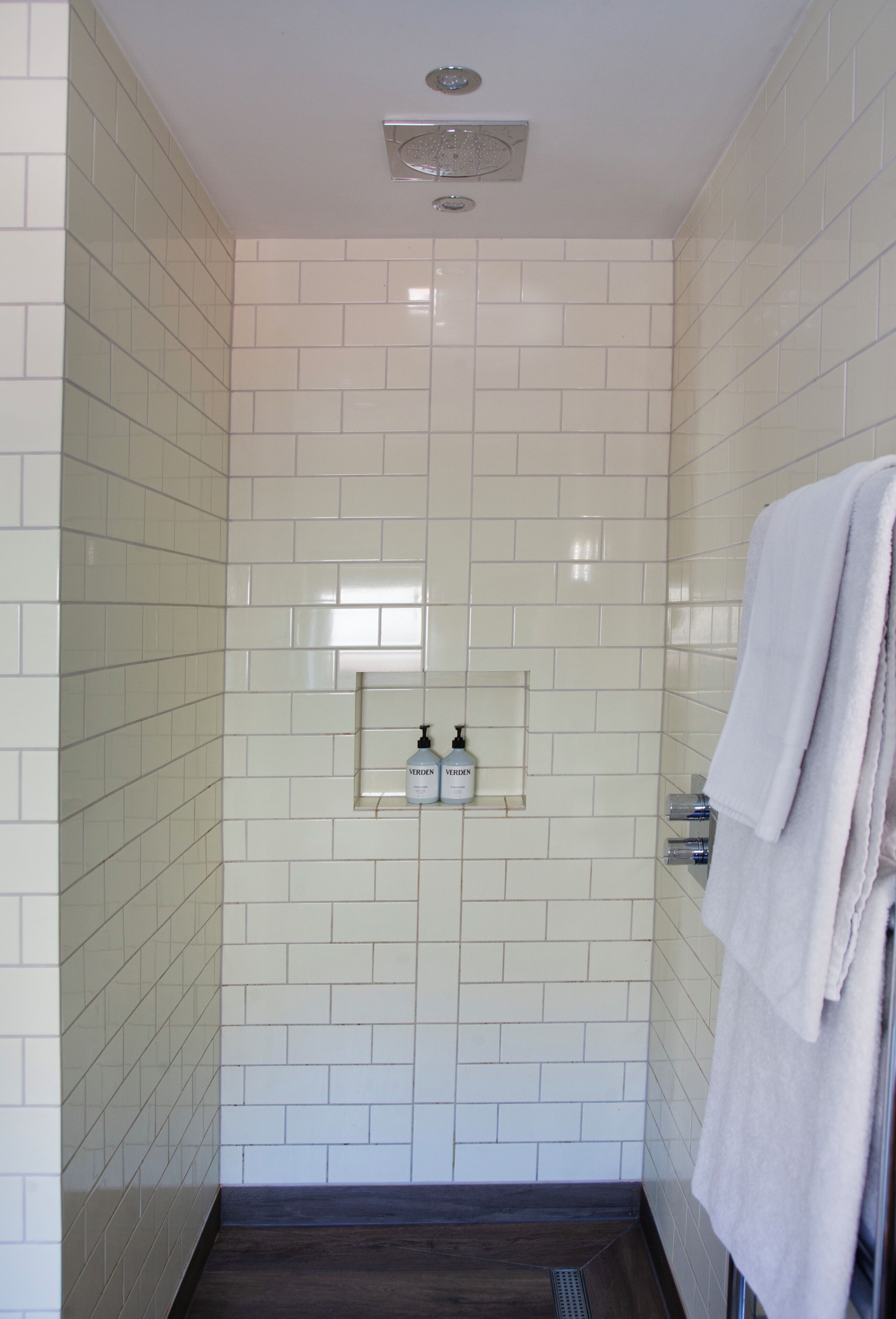 A bathroom with white tiled walls, a showerhead, a shelf with toiletries, and a towel rack.