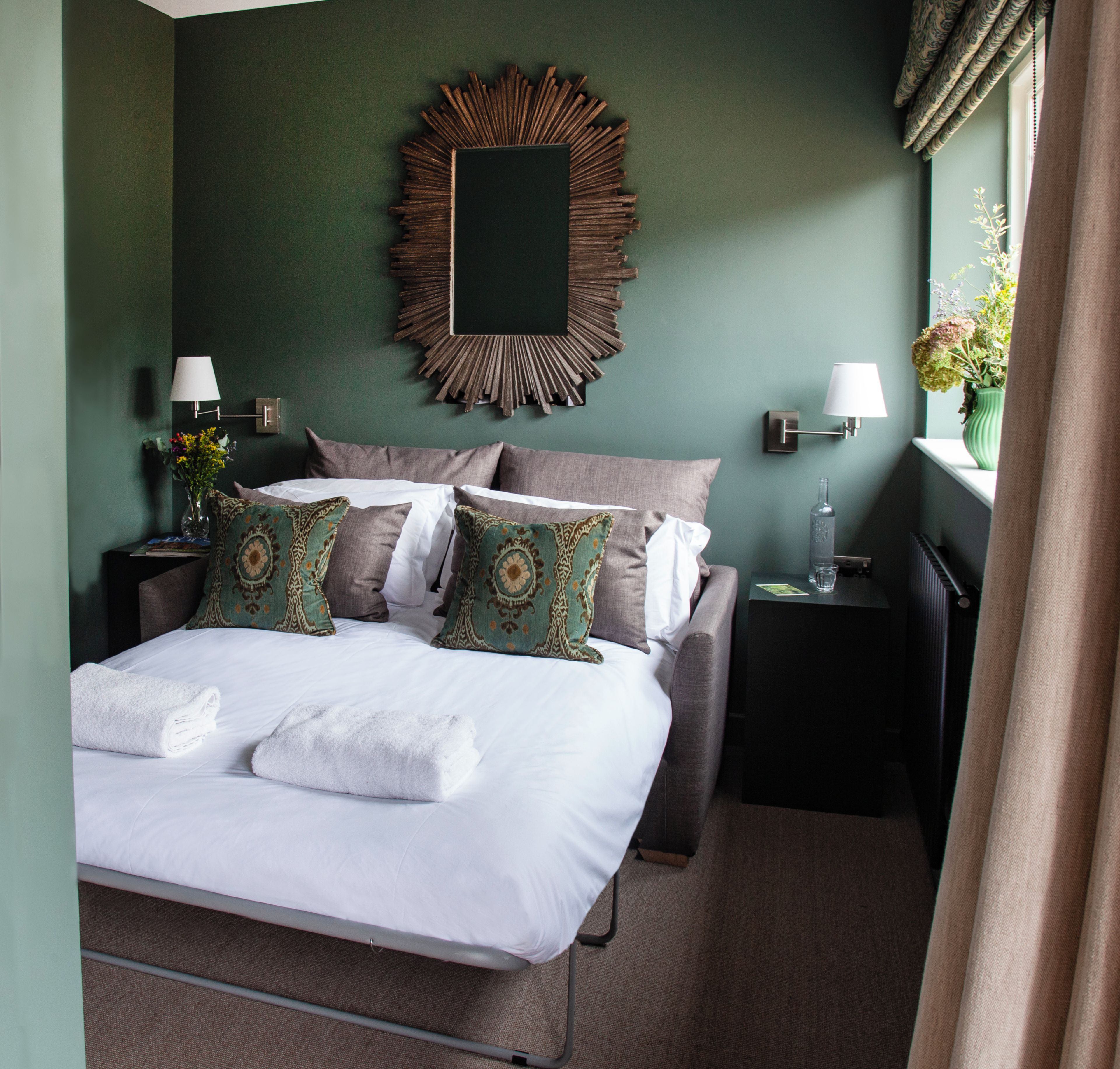 Una cama bien hecha con sábanas blancas y almohadones decorativos, un espejo montado en la pared y una mesita de noche con una lámpara y un florero.