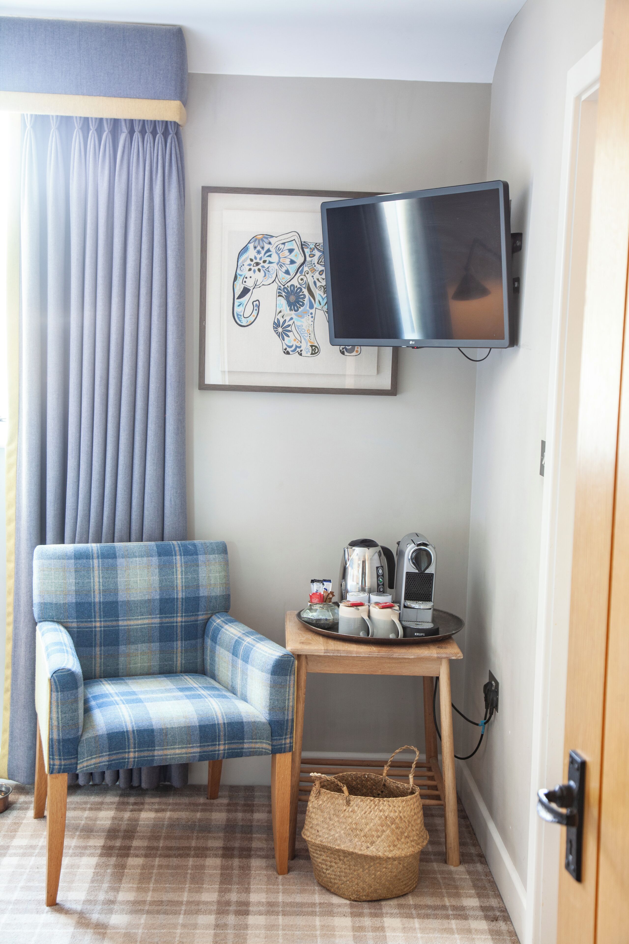 A corner room with a plaid chair, a small table with a kettle, a basket, and a framed elephant picture.