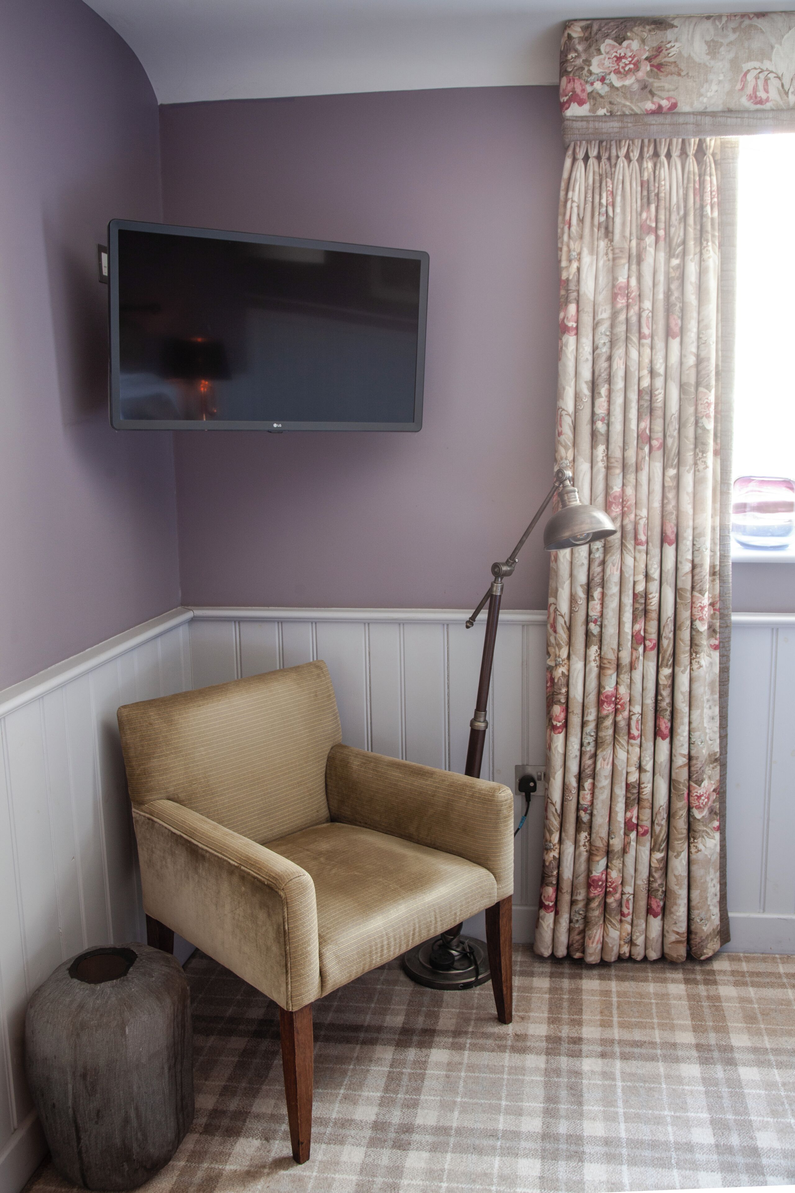Una habitación con un televisor de pantalla plana montado en la pared, un sillón color beige, una lámpara de pie y una alfombra a cuadros.