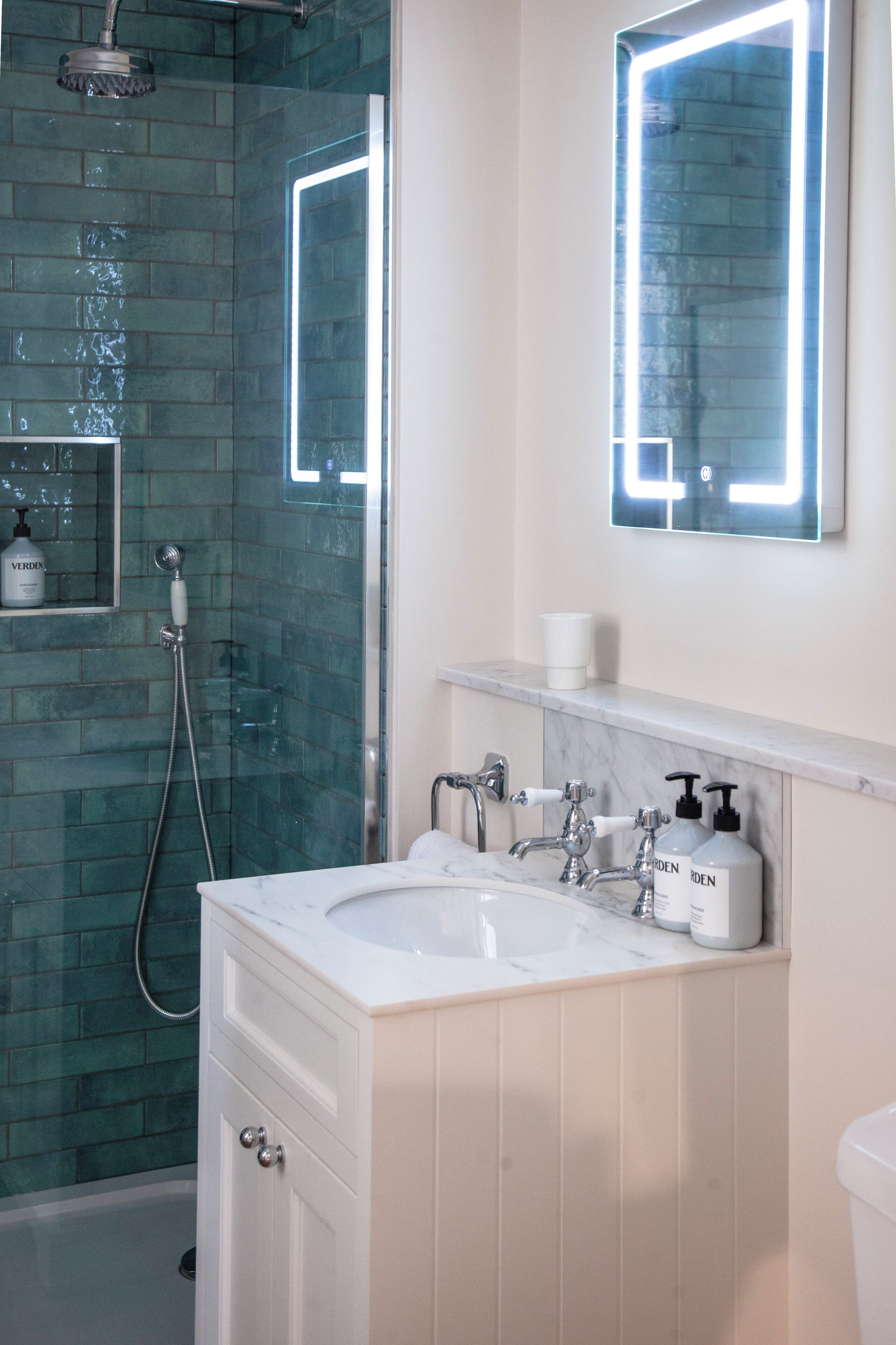 A bathroom with a glass shower enclosure, a white sink with two faucets, and a mirror with integrated lighting.
