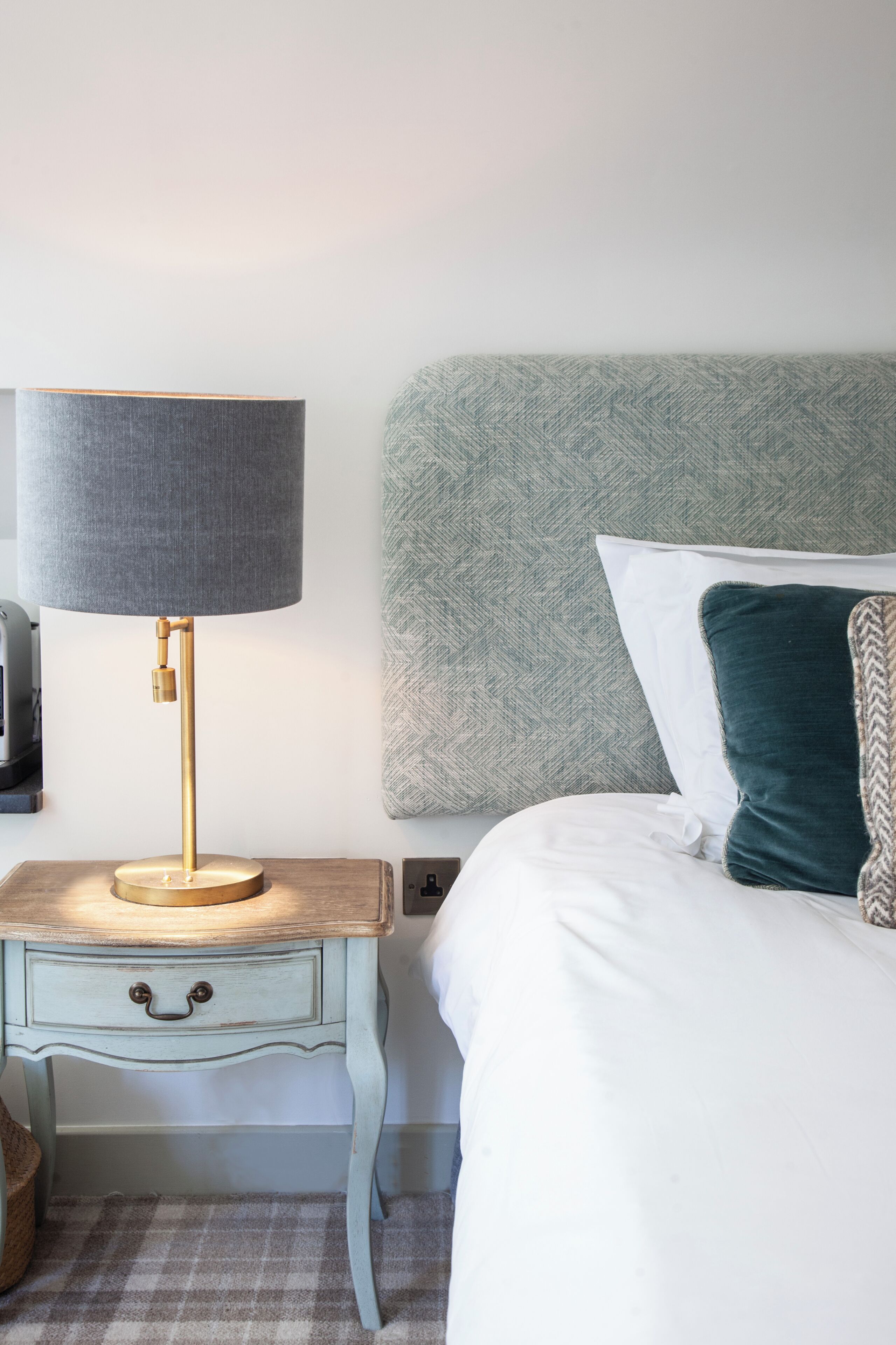 A neatly made bed with a headboard, a bedside table with a lamp, and a small drawer.