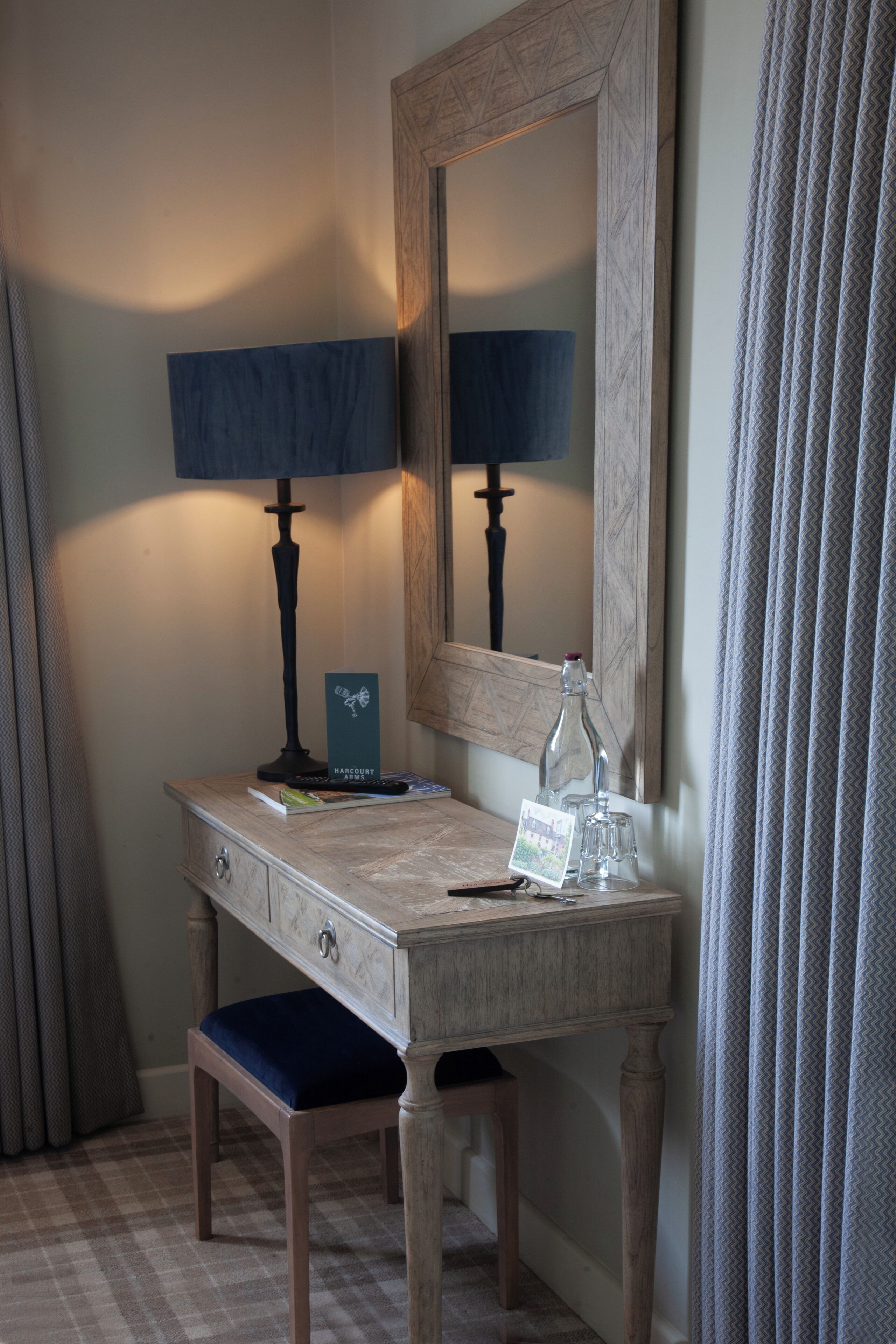 A wooden desk with a lamp, a mirror, a chair, and a small table with a bottle and a card.