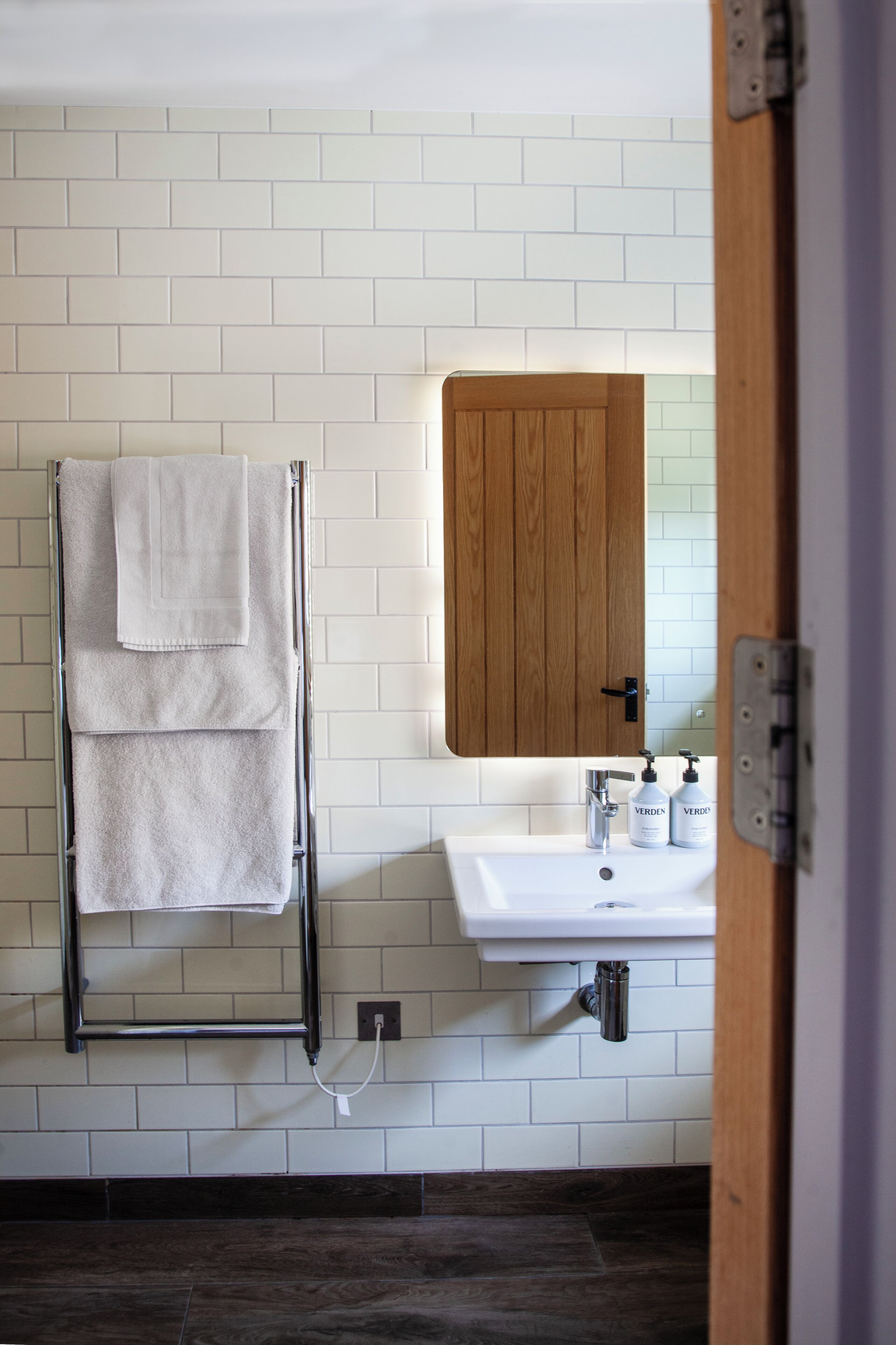 A bathroom with a white tiled wall, a wooden door, a towel rack with two towels, a sink with two bottles, and a visible plumbing pipe.