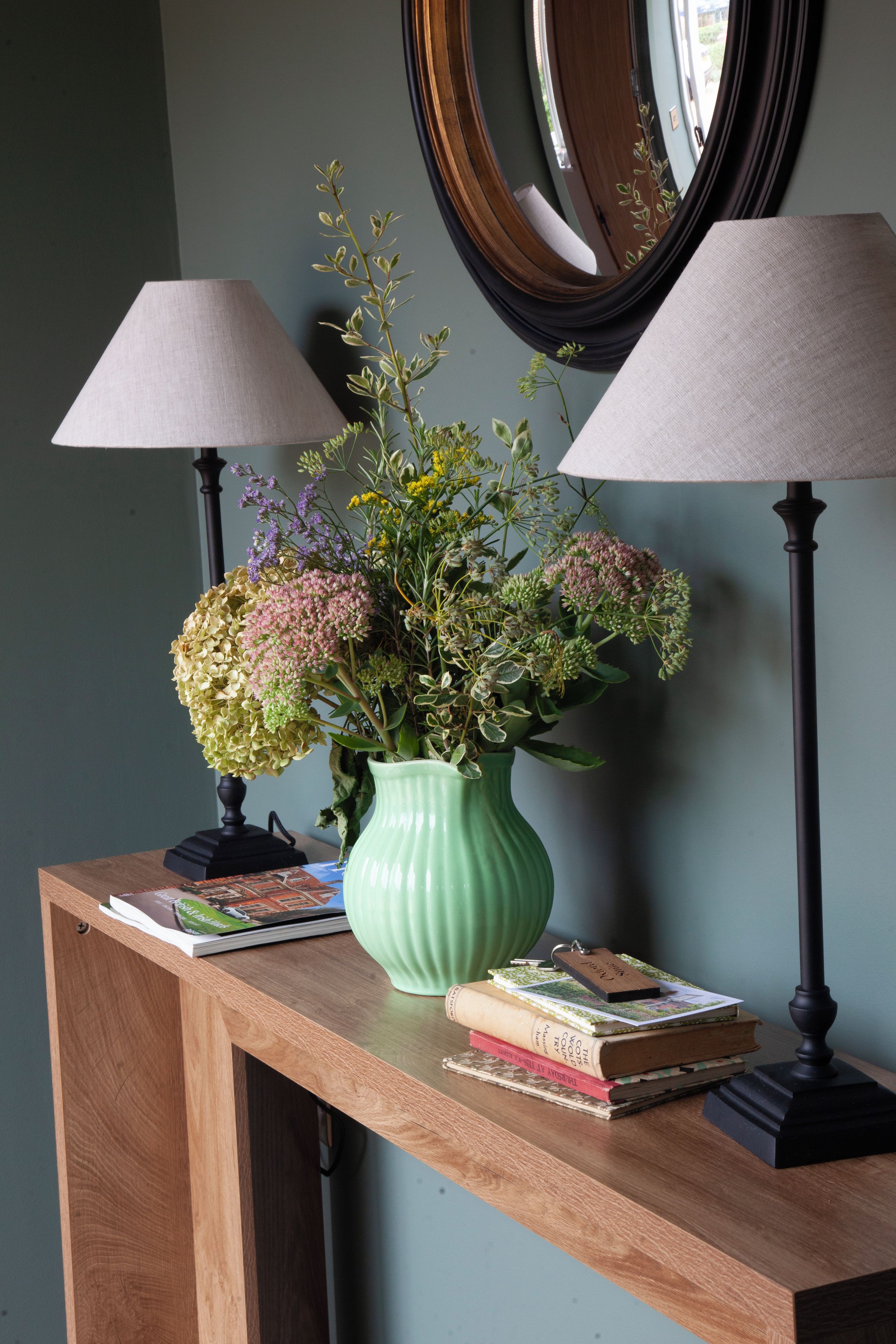Een houten console tafel met een groene vaas vol bloemen, twee lampen en een stapel boeken.