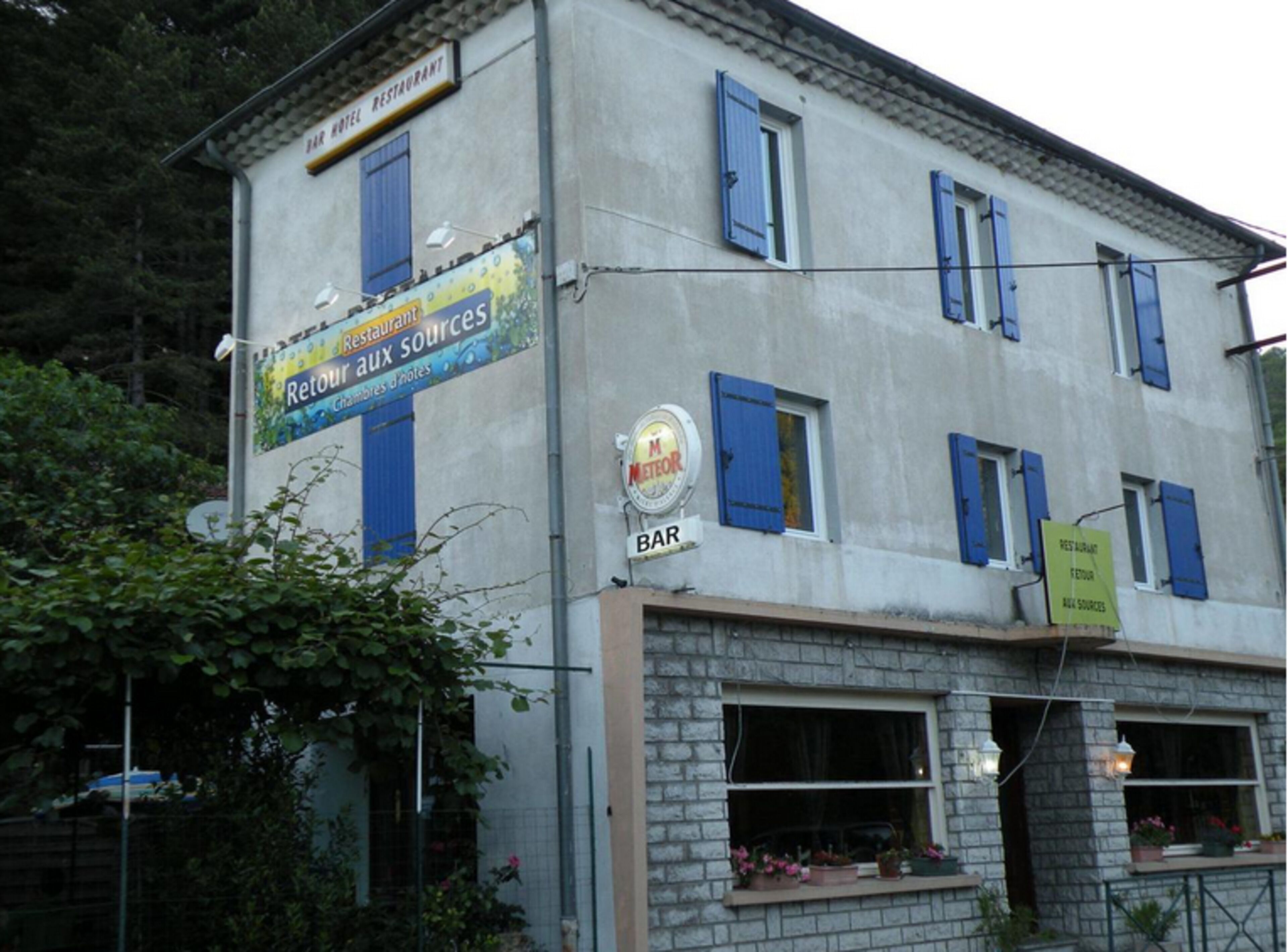 Un bâtiment avec une enseigne indiquant un restaurant et un bar, doté d’une fenêtre à volets bleus et d’une autre enseigne signalant un bar à cocktails.
