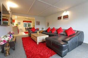 A living room with a sofa, coffee table, and a red rug. There is a kitchen area in the background. - Groovy on Grey - Spa Pool, Central, Sleeps 8 (Rotorua)