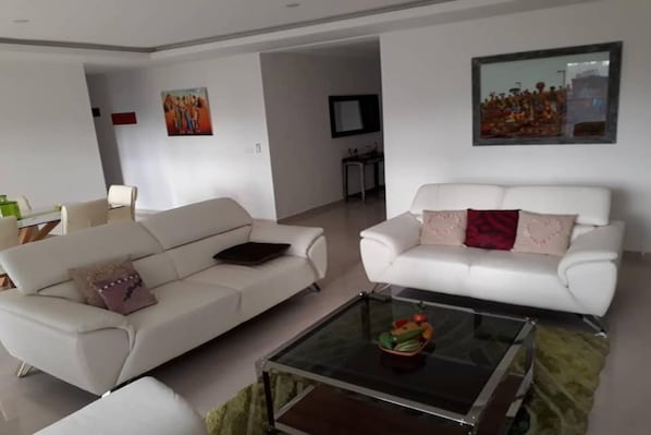 A living room with two white sofas, a glass coffee table, and a dining area in the background.