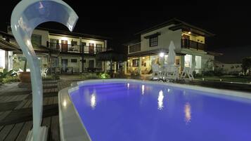 Una piscina con una estructura blanca curva en primer plano, rodeada por una terraza de madera. Al fondo, casas de dos pisos con balcones y zonas de estar al aire libre.