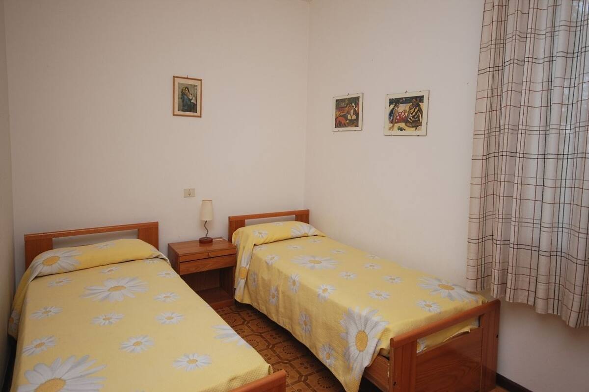 Una habitación pequeña y sencilla con dos camas individuales, una mesita de noche de madera, una lámpara y dos cuadros en la pared.