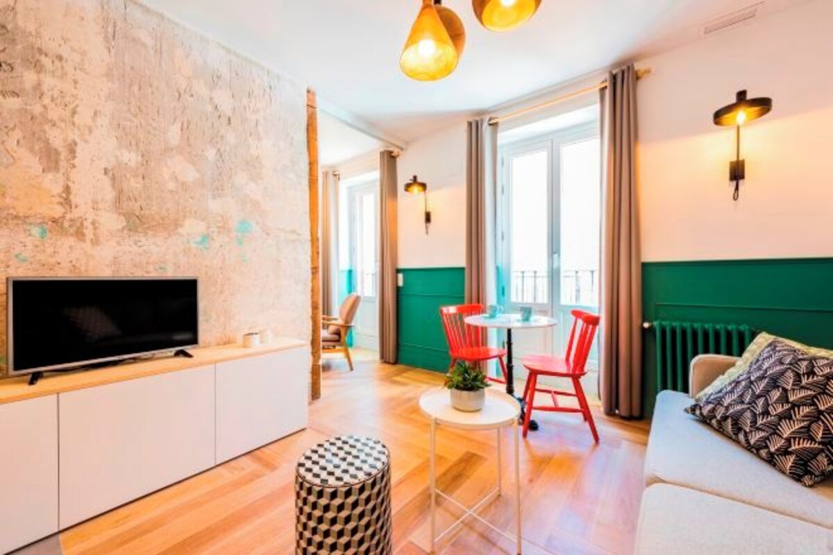 Una sala moderna con televisor de pantalla plana, una mesa de comedor redonda con sillas rojas, un reposapiés estampado y un sofá con cojines decorados.