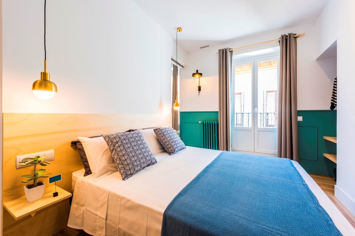 Habitación de hotel con cama, mesita de noche, planta en maceta y vistas al exterior.