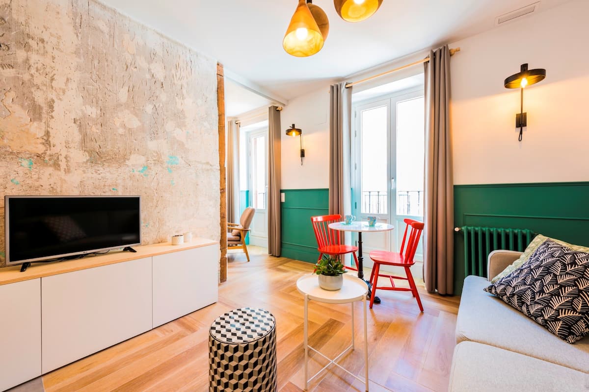 Una sala moderna con un televisor de pantalla plana, una mesa de comedor redonda, sillas rojas, un reposapiés estampado y un sofá con cojines decorativos.