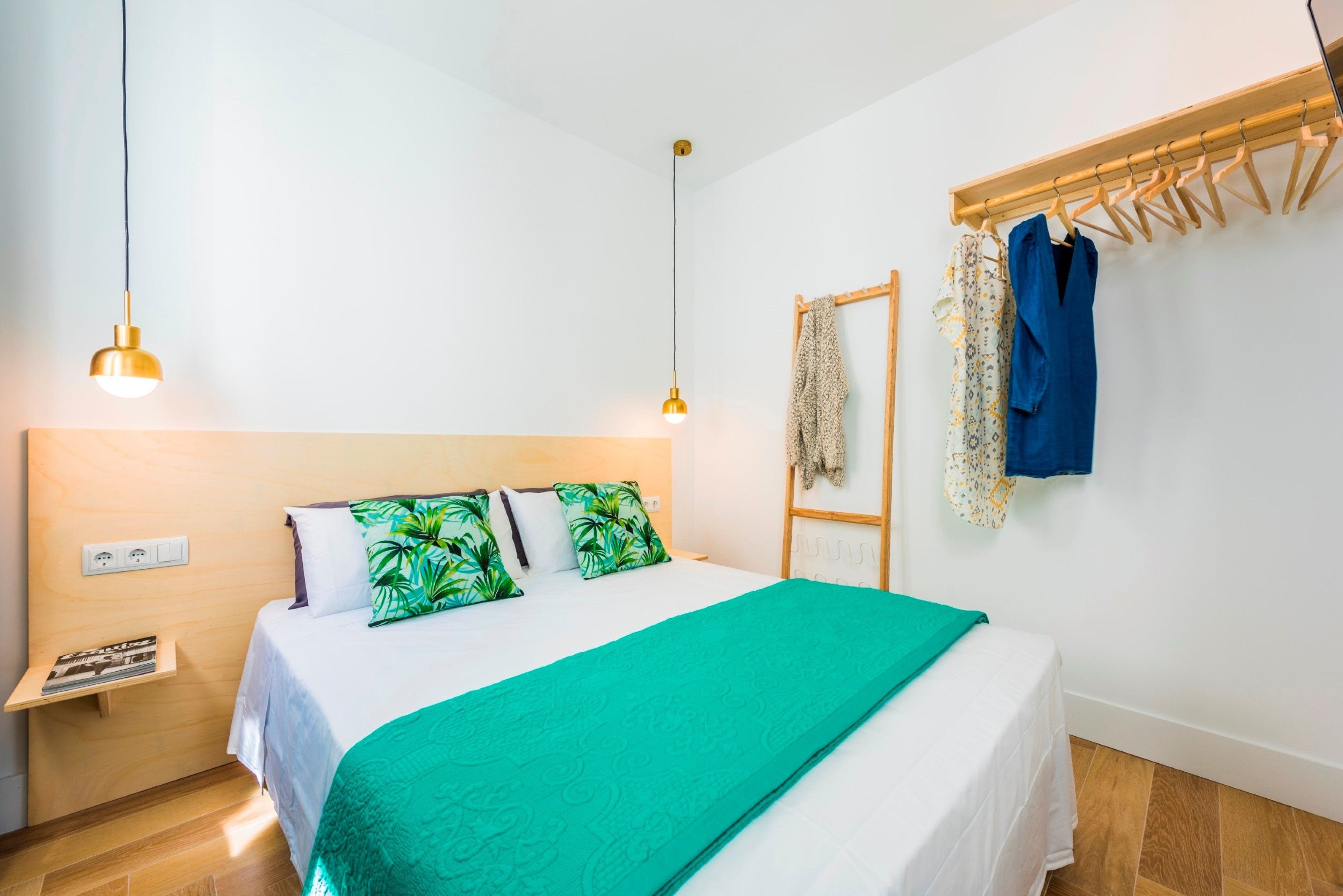 A neatly made bed with a green blanket, a wooden headboard, and a wooden shelf with a book.