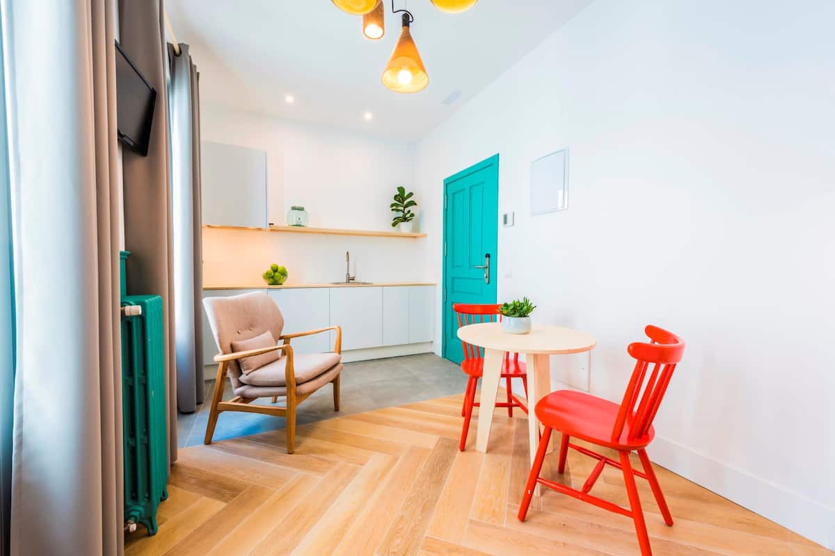 Cocina moderna con armarios blancos, una puerta verde azulada y un pequeño comedor con una mesa y sillas.
